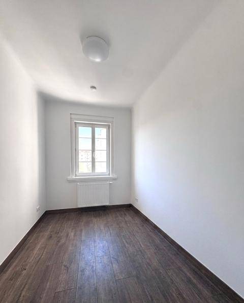 Kompaktes, helles Zimmer mit dunklem Holzboden und einem Fenster, ideal als Kinder- oder Gästezimmer.