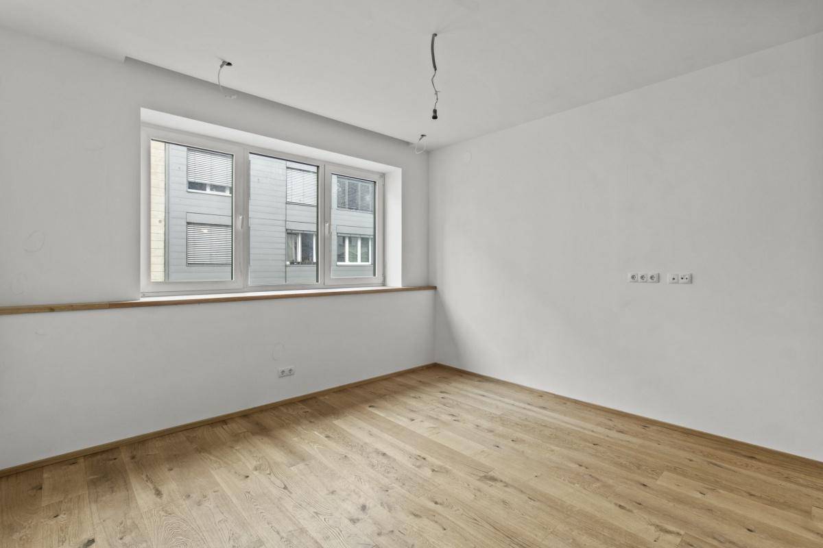 Helles Zimmer mit Holzboden und einem Fenster, das den Blick auf die Nachbargebäude ermöglicht.