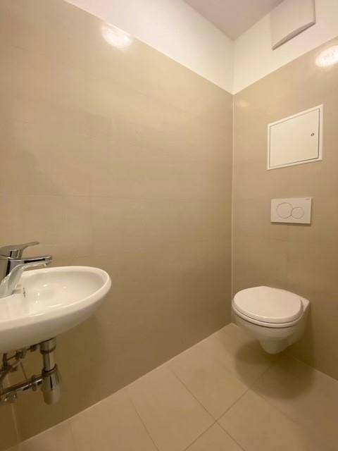 Separates WC mit wandmontierter Toilette, kleinem Waschbecken und hellen Fliesen.