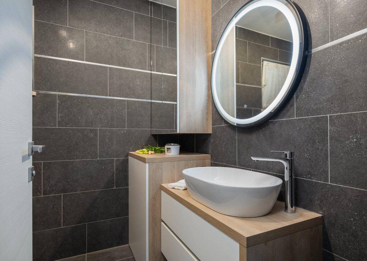 Modernes Badezimmer mit dunklen Fliesen, ovalem Waschbecken und beleuchtetem Spiegel.