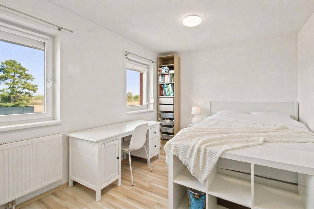 Helles Schlafzimmer mit weißem Bett, Schreibtisch und Blick ins Grüne durch große Fenster.