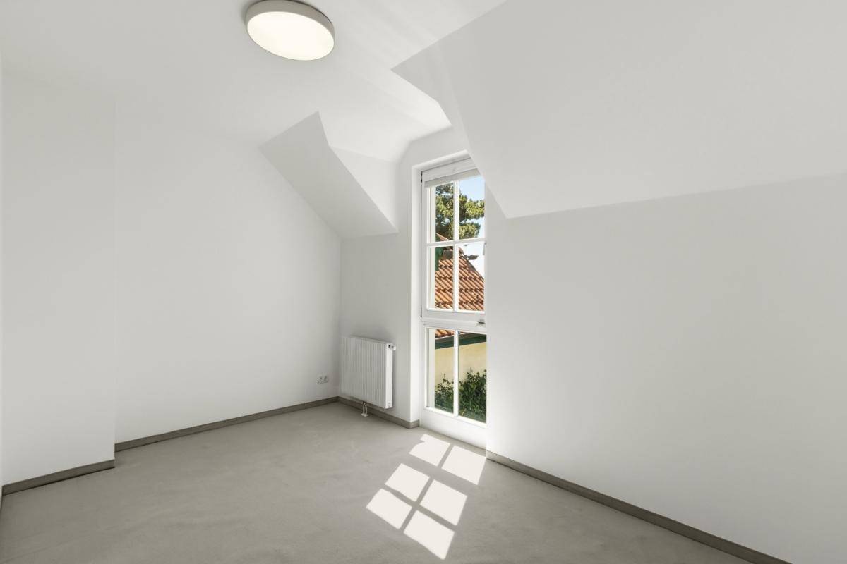 Ruhiges Zimmer mit Teppichboden, weiß gestrichenen Wänden und einem Fenster mit Blick ins Grüne.