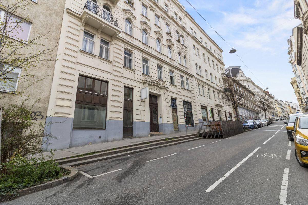 Gesamtansicht eines historischen Gebäudes an einer Straße mit parkenden Autos und Bäumen im städtischen Umfeld.