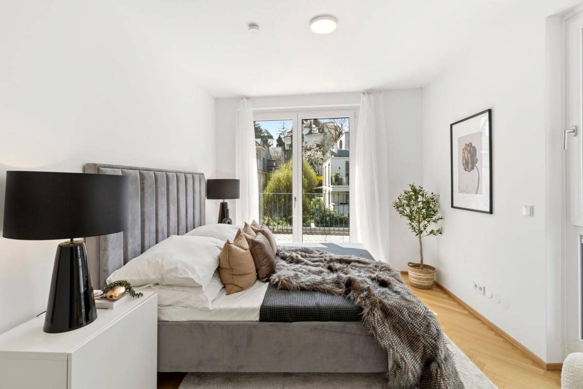 Modernes Schlafzimmer mit grauem Polsterbett, Parkettboden und Balkonzugang mit Blick ins Grüne.
