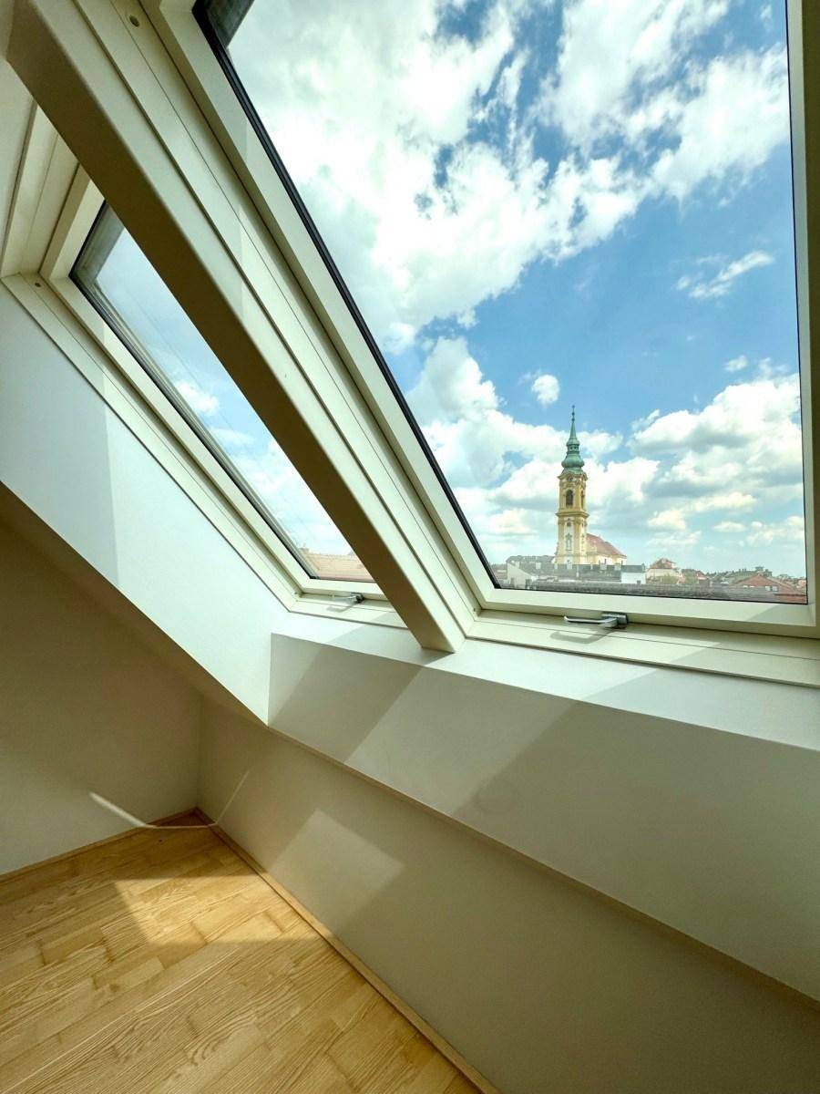 Helles Zimmer mit Holzparkett und einem Dachfenster, das einen weiten Blick auf die Stadt und einen Kirchturm bietet.