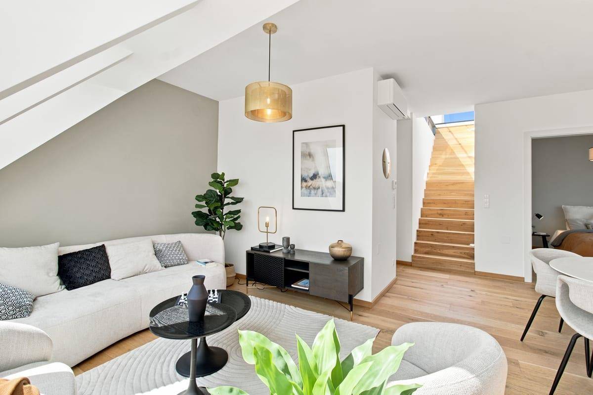 Helles Wohnzimmer mit Sofa, Sesseln und Holztreppe, die zur oberen Ebene führt.