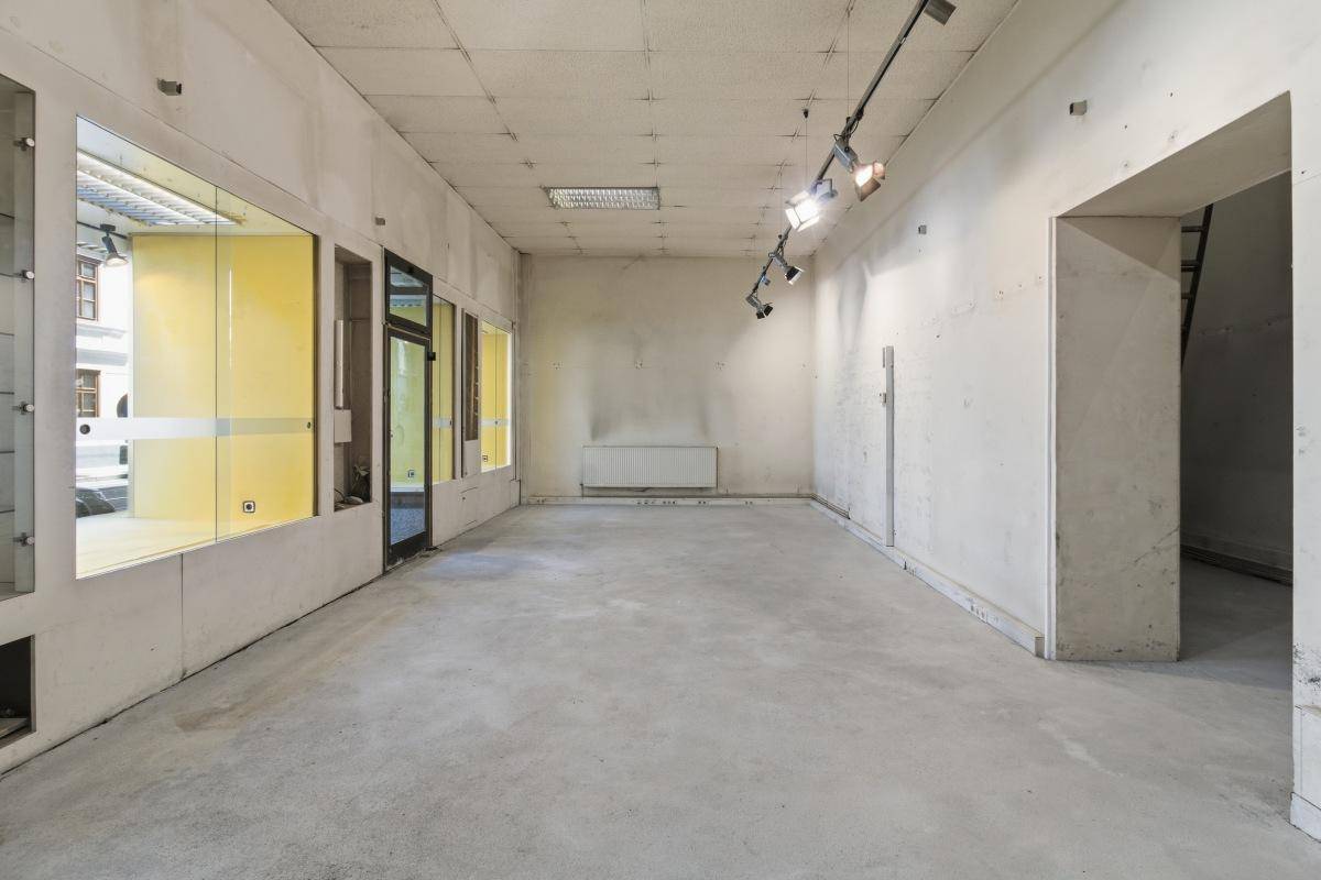 Langer, leerer Ladenraum mit Betonboden, Schaufenstern und einem Durchgang zu einem hinteren Bereich.