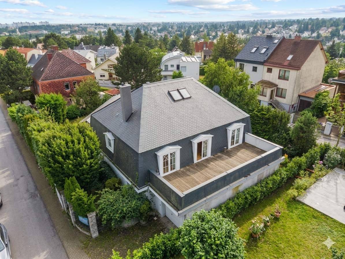Vogelperspektive eines Wohnhauses mit dunklem Mansarddach, großer Holzterrasse und umgebender Hecke.