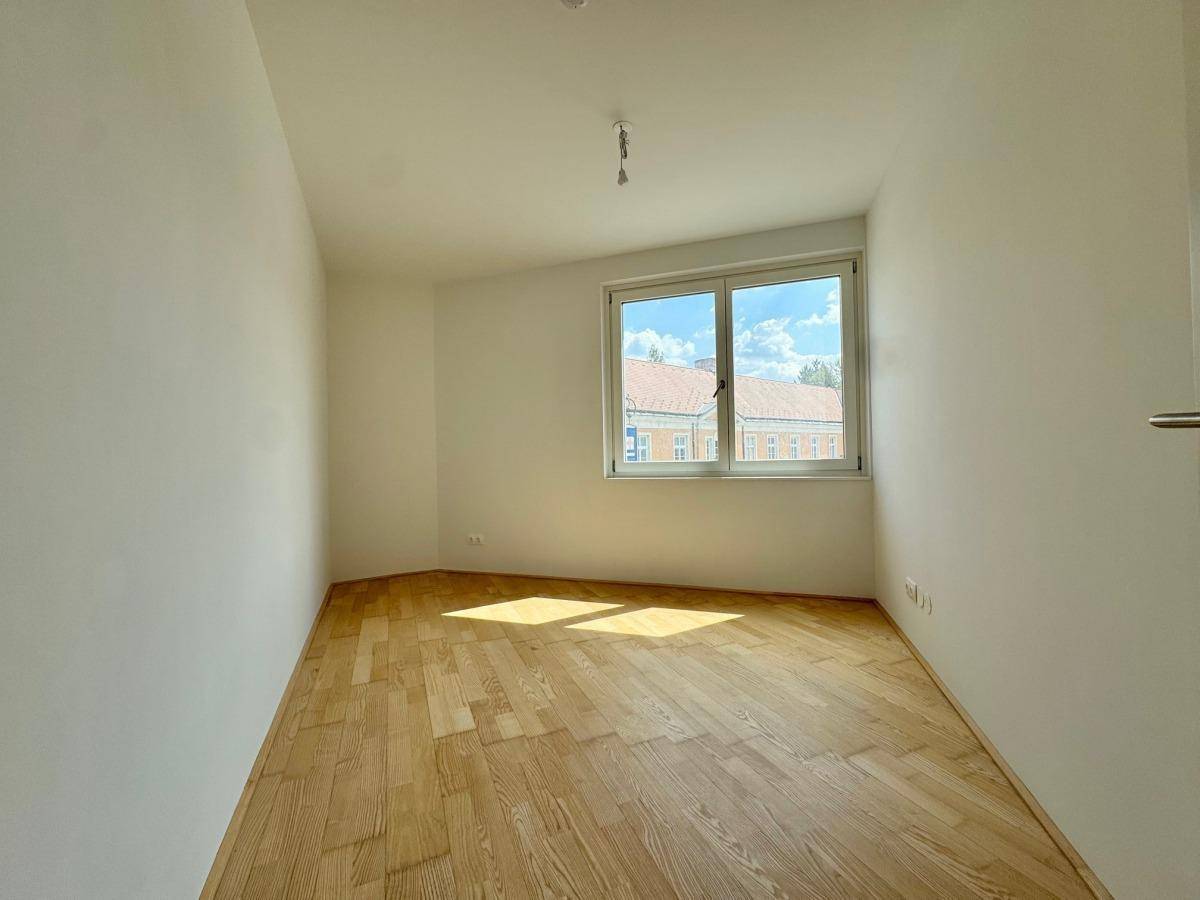 Helles Schlafzimmer mit Parkettboden und großem Fenster, das viel Sonnenlicht hereinlässt.