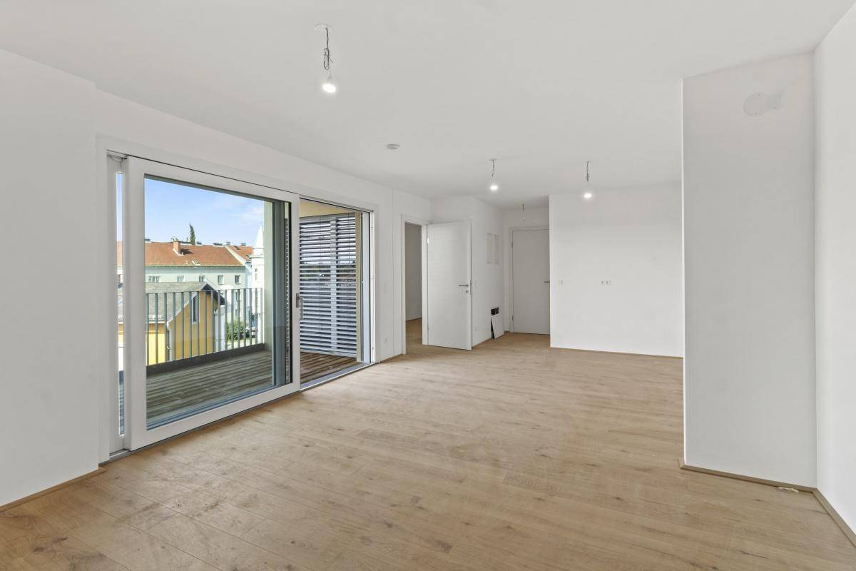 Großer, heller Wohnbereich mit Holzboden und Zugang zu einem Balkon mit Jalousien.