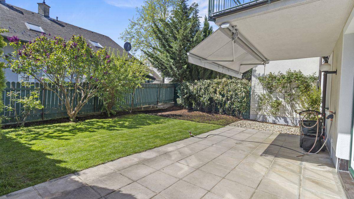 Geräumige Terrasse mit Rasenfläche, umgeben von Bäumen und einem ausfahrbaren Sonnenschutz.