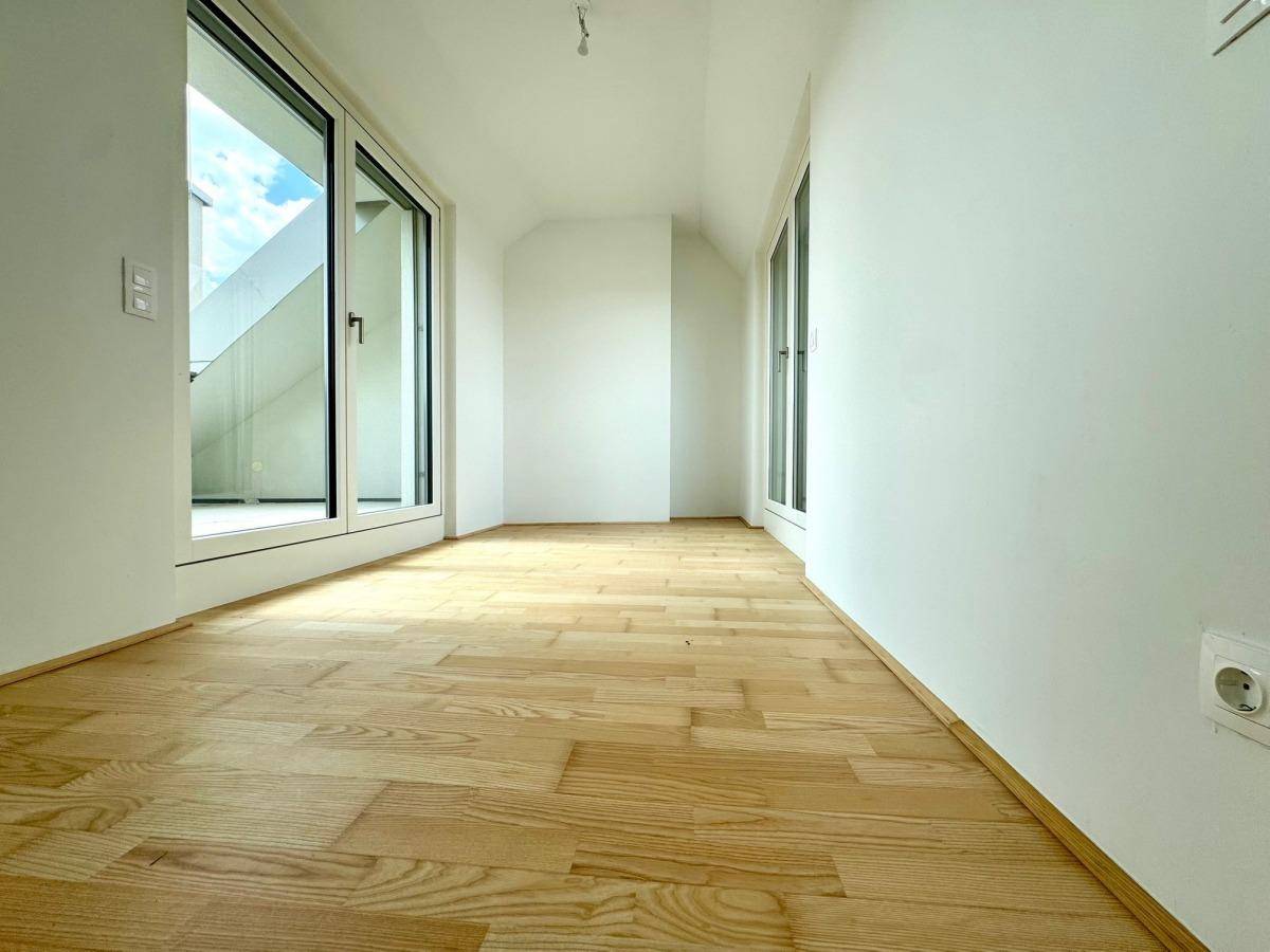 Heller Raum mit Holzboden und großen Glasfenstern, die Zugang zu einem Außenbereich bieten.