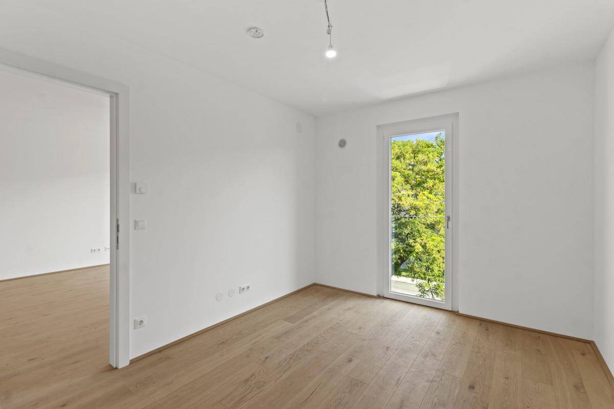 Helles Schlafzimmer mit Parkettboden und einem hohen Fenster, das viel Tageslicht hereinlässt.