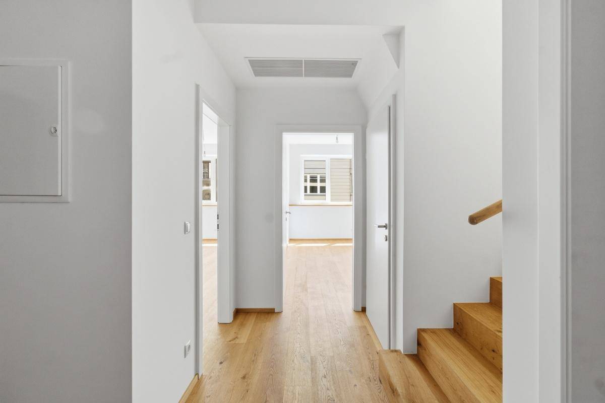 Heller Flur mit Holzboden und weißen Wänden, der zu verschiedenen Zimmern und einer Treppe führt.