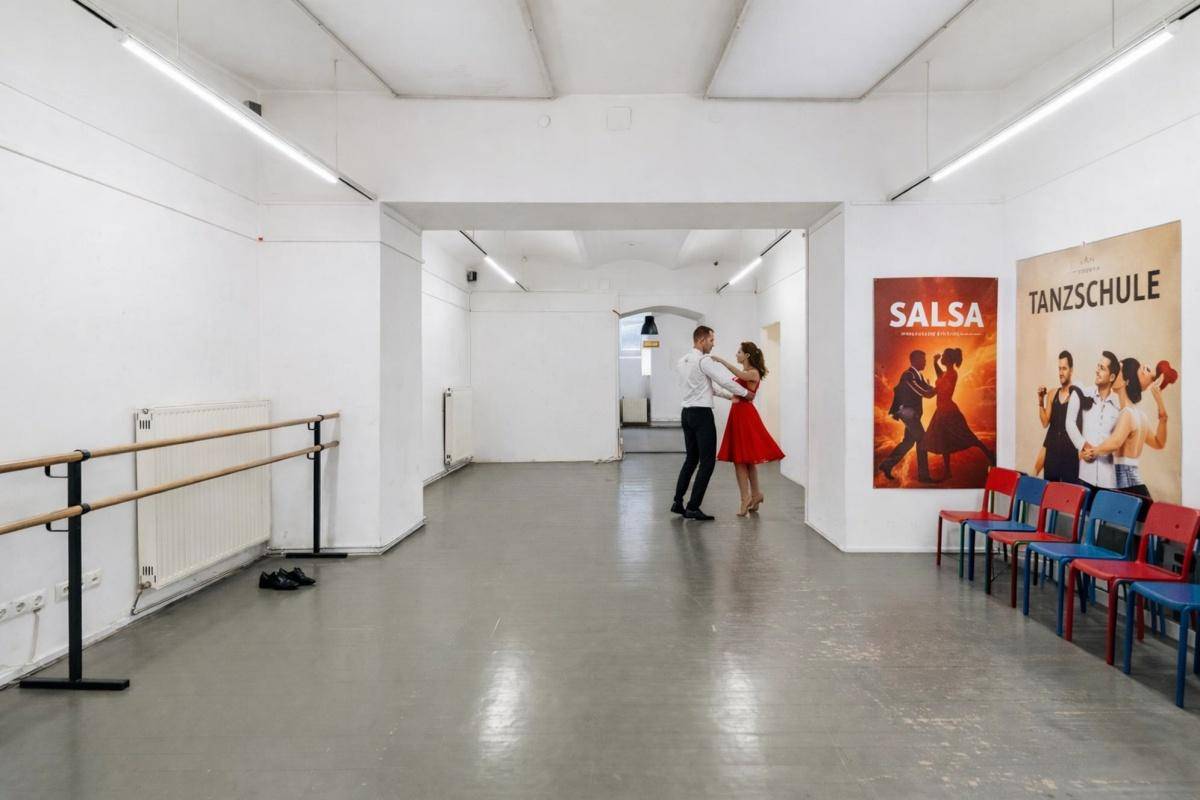 Großer Tanzsaal mit Ballettstangen, Plakaten und einem Paar beim Tanzen, mit hohen Decken.