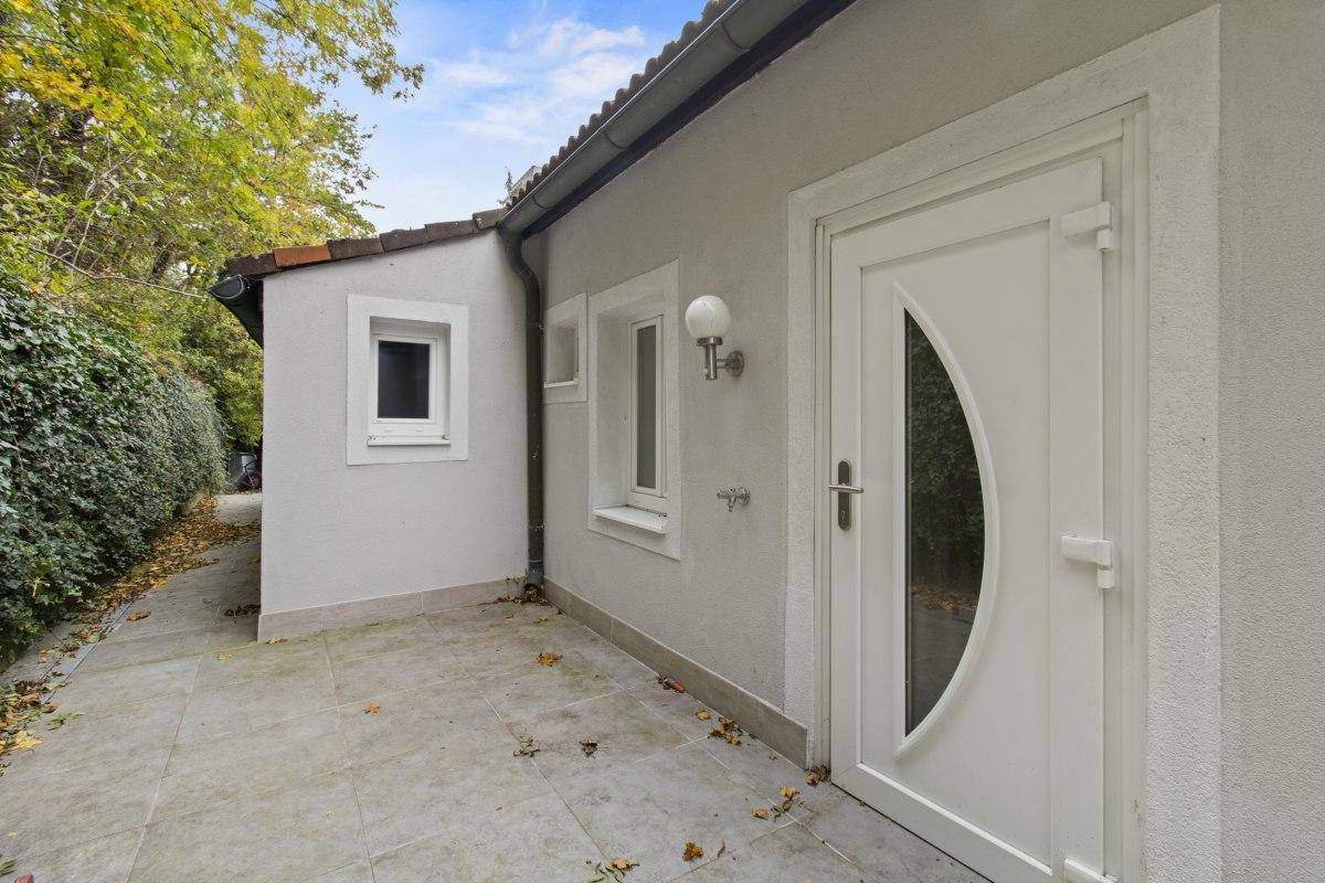 Seitlicher Eingangsbereich des Gebäudes mit weißer Tür und gepflastertem Vorplatz.
