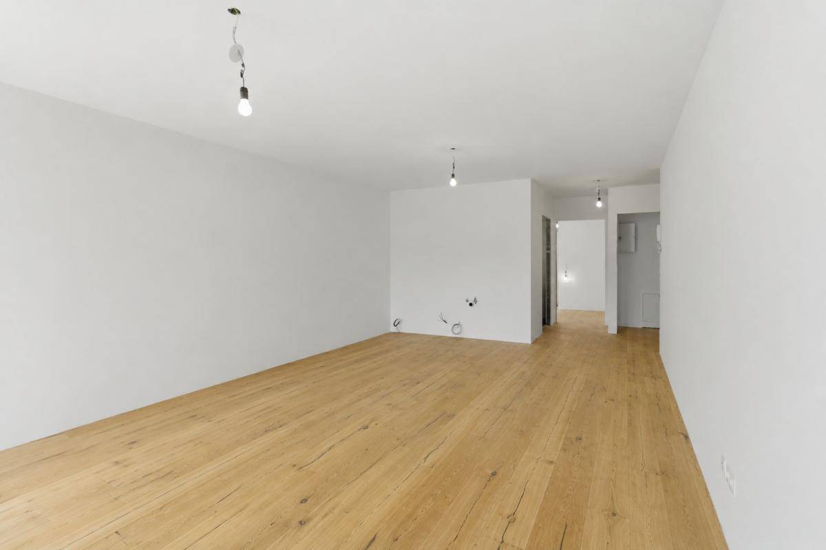 Geräumiger Raum mit Holzboden und weiß gestrichenen Wänden, offen zum Flurbereich.