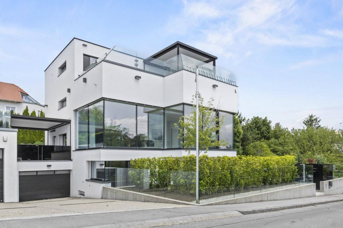 Moderne weiße Villa mit großen Glasfronten, klaren Linien und gepflegter Hecke an der Straße.