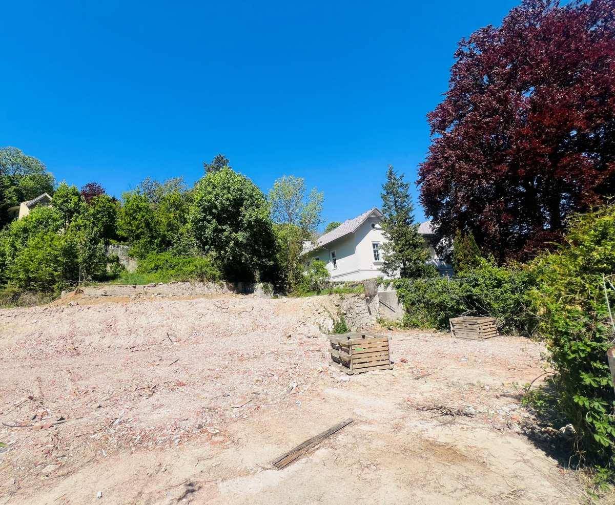 Ein sonniges Baugrundstück mit Blick auf ein bestehendes Haus und grüne Hügel im Hintergrund.