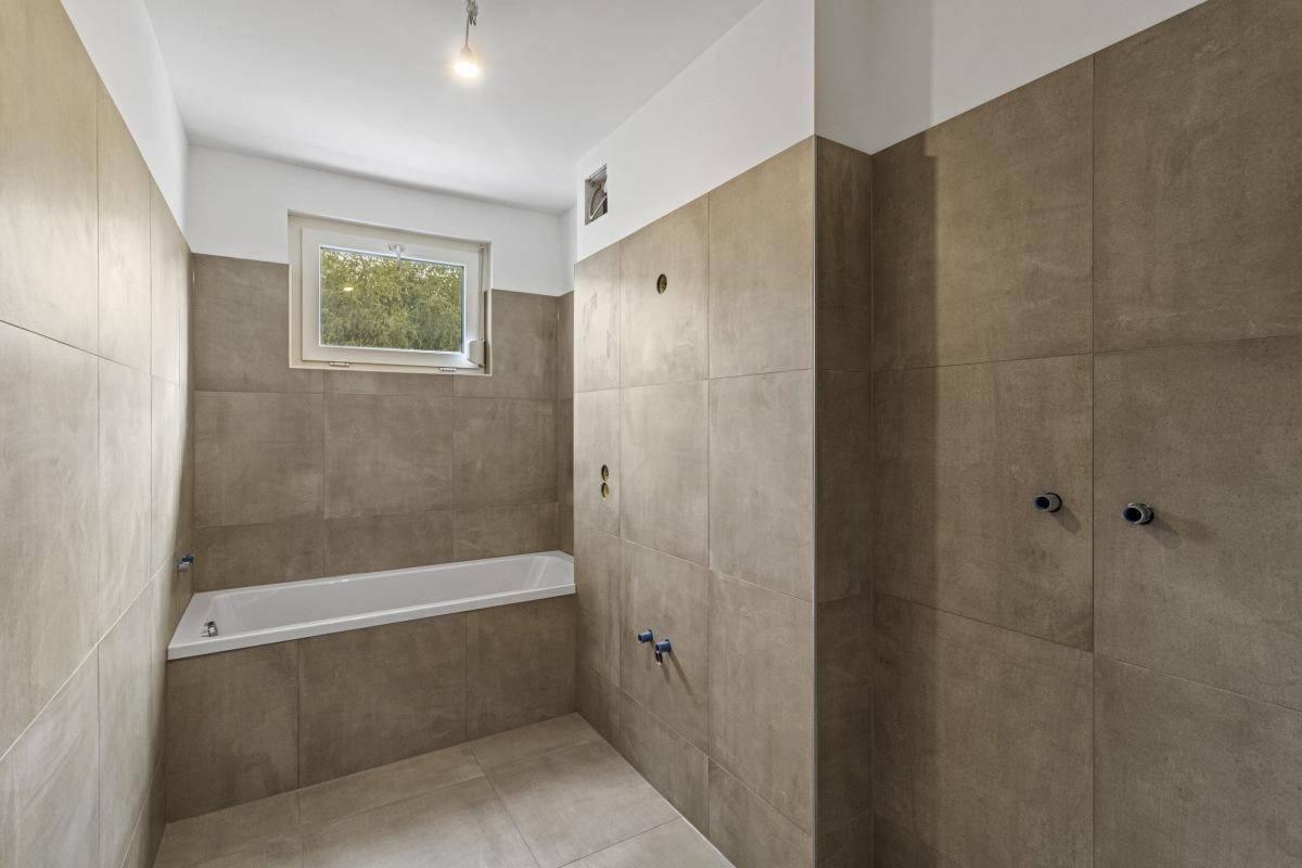 Unfertiges Badezimmer mit braunen Fliesen, eingebauter Badewanne und Fenster für Tageslicht.