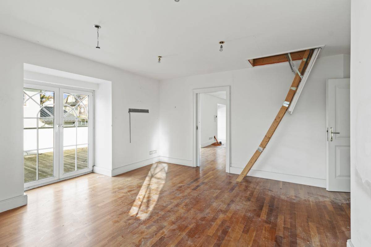 Leeres Zimmer mit Holzboden, großen Fenstern und einer Leiter zum Dachboden oder einer oberen Ebene.