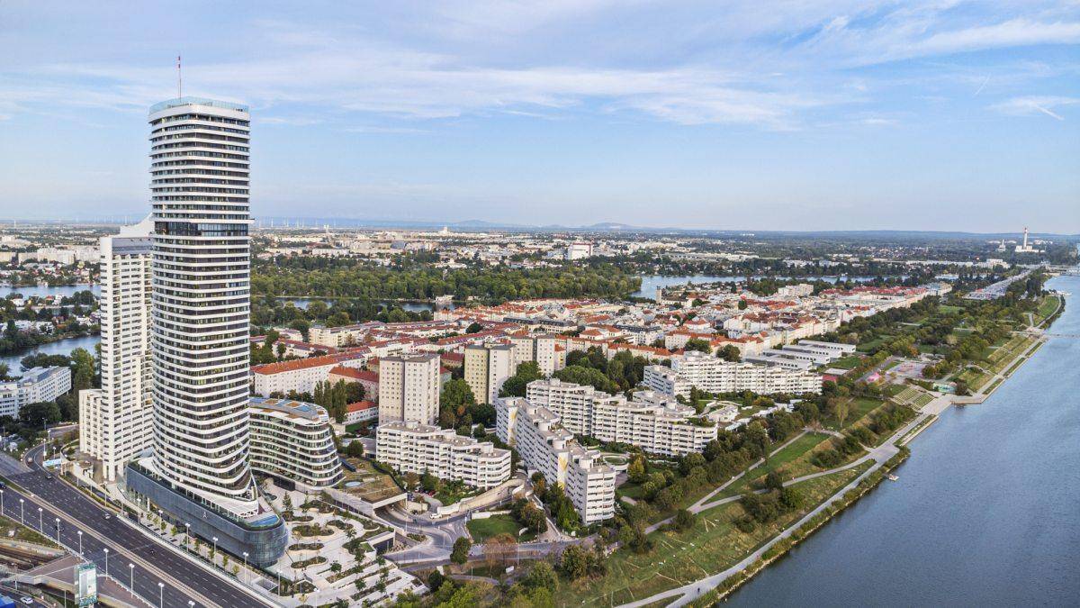Luftaufnahme der Stadt mit einem markanten Hochhaus am Flussufer und weitläufiger Bebauung.