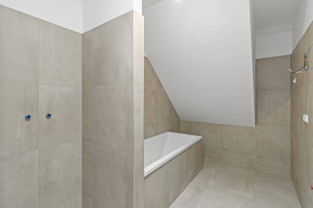 Badezimmer im Bau mit hellgrauen Fliesen, eingebauter Badewanne und Dachschräge.