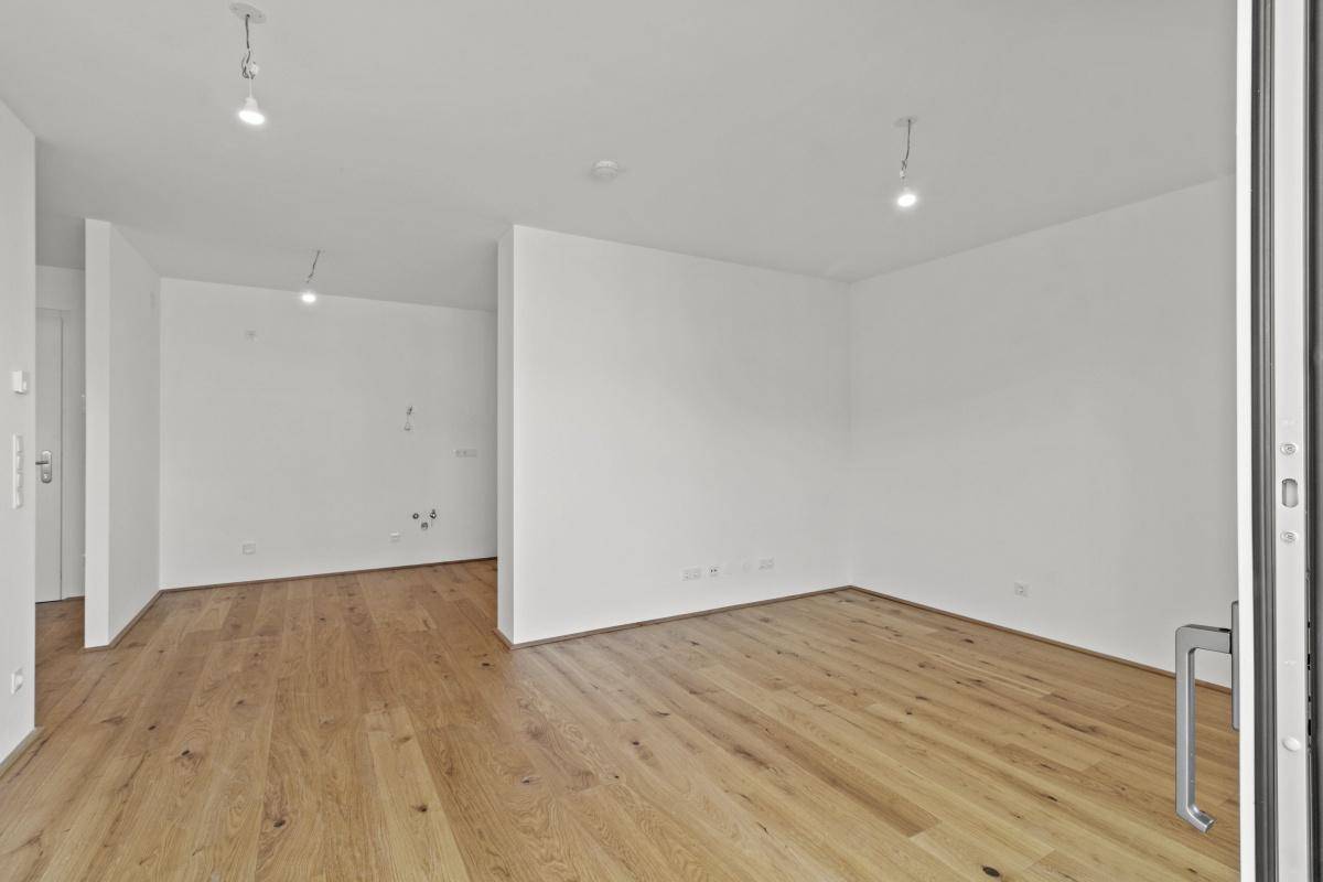 Unmöbliertes Wohnzimmer mit hellem Holzboden und viel Tageslicht durch die Balkontür.