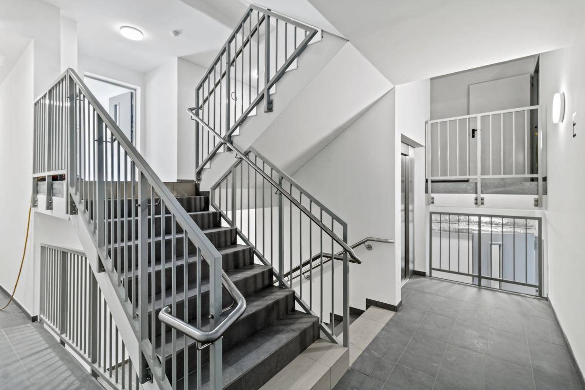 Treppenhaus und Aufzug in einem modernen Gebäude, mit Metallgeländern und Fliesenboden.