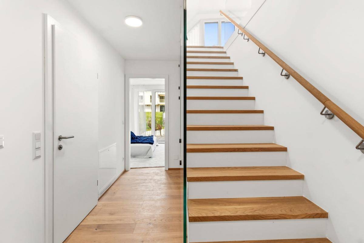 Heller Flur mit Holztreppe, Glasgeländer und Blick auf den Außenbereich.