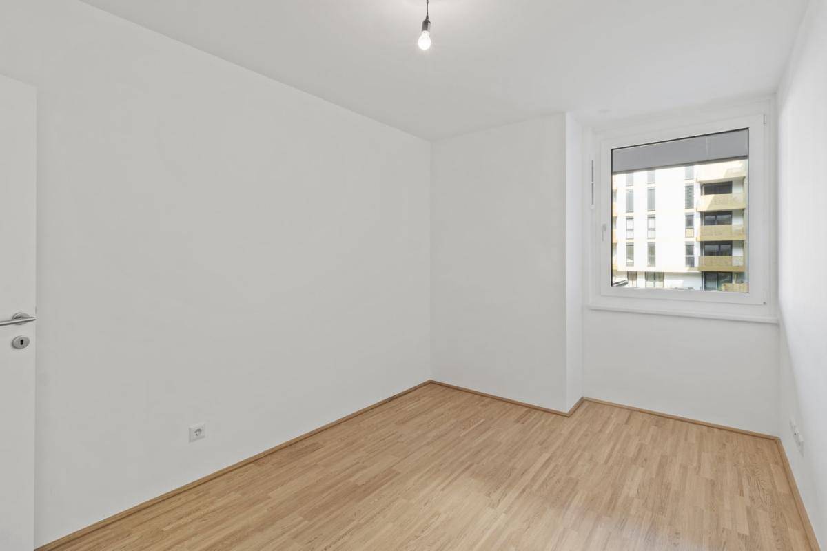 Leeres Schlafzimmer mit Holzboden und einem großen Fenster, das viel Tageslicht hereinlässt.