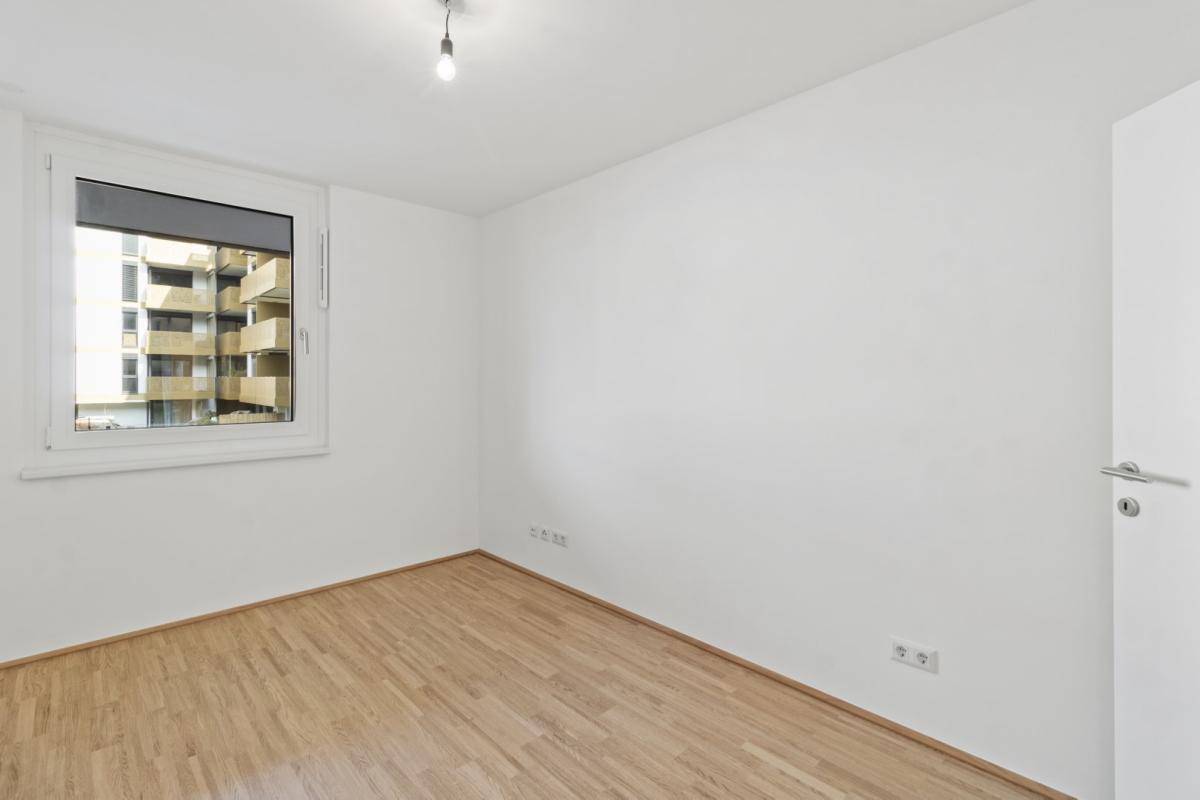 Helles, unmöbliertes Schlafzimmer mit Holzboden und einem Fenster mit Blick auf andere Gebäude.