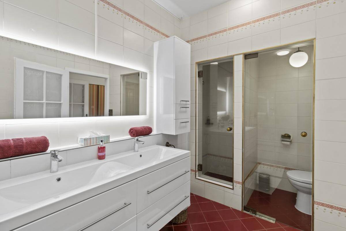 Helles Badezimmer mit Doppelwaschbecken, Duschkabine und roten Bodenfliesen.