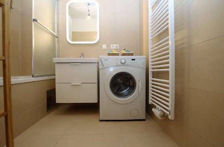 Helles Badezimmer mit beigefarbenen Fliesen, Waschbecken, Waschmaschine und Badewanne.