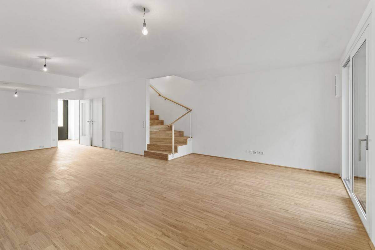 Großes, leeres Wohnzimmer mit Holzboden und einer eleganten Holztreppe zum Obergeschoss.