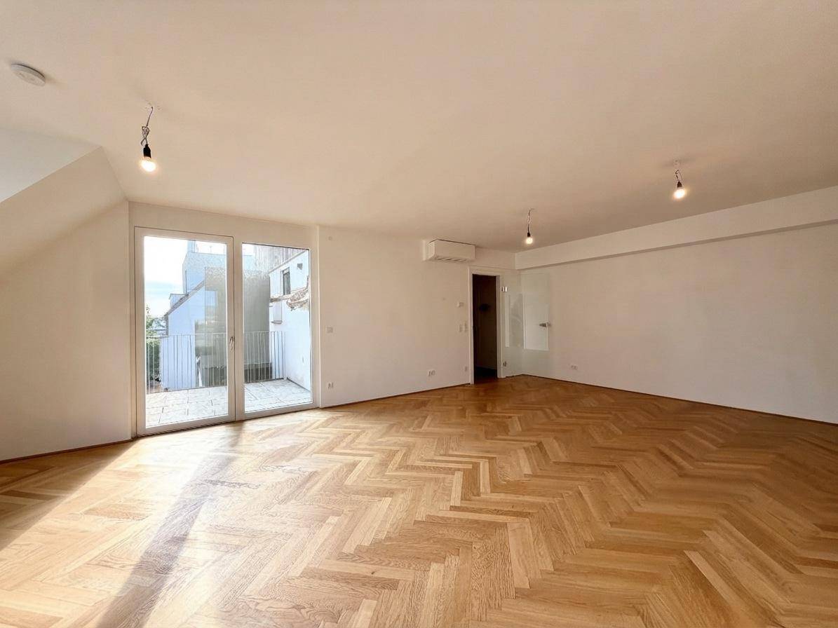 Geräumiger, leerer Wohnbereich mit Fischgrätparkett und großen Fenstern zum Balkon.