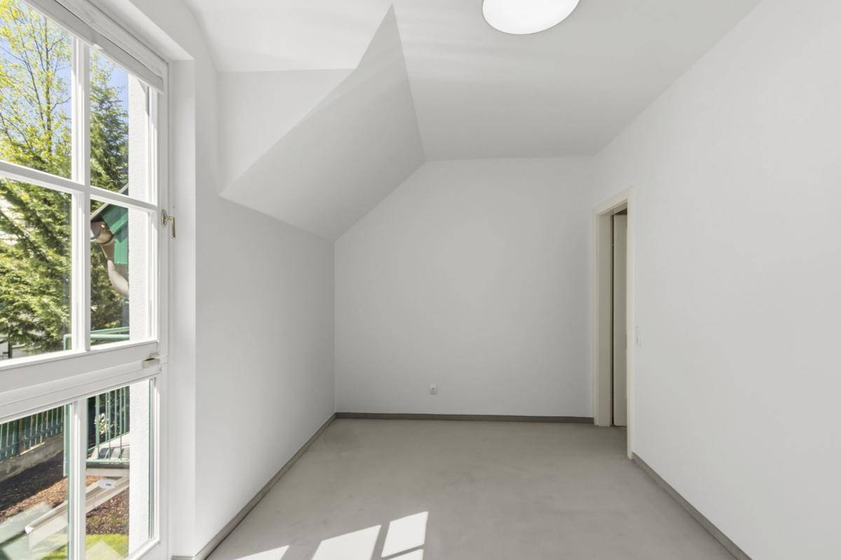 Helles Zimmer mit Teppichboden, schrägen Wänden und einem Fenster, das viel Tageslicht hereinlässt.