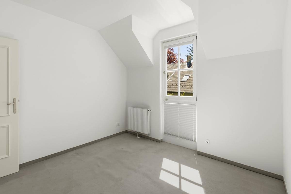 Helles Zimmer mit Teppichboden, schrägen Wänden und einem Fenster mit Blick auf die Dächer.