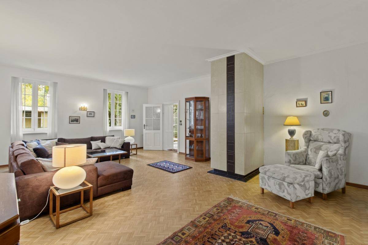 Großzügiges Wohnzimmer mit Parkettboden, Kachelofen und mehreren Fenstern, die viel Tageslicht hereinlassen.