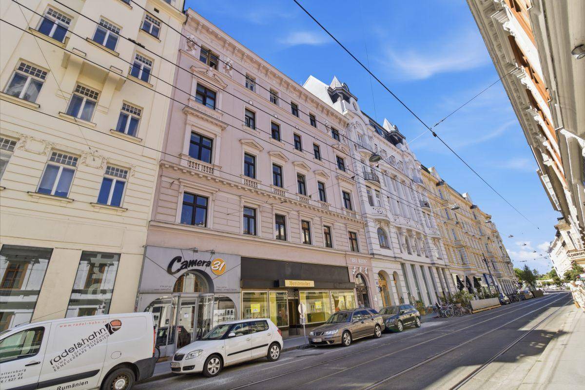Gesamtansicht des historischen Gebäudes mit Geschäftsfassaden und der belebten Straße an einem sonnigen Tag.
