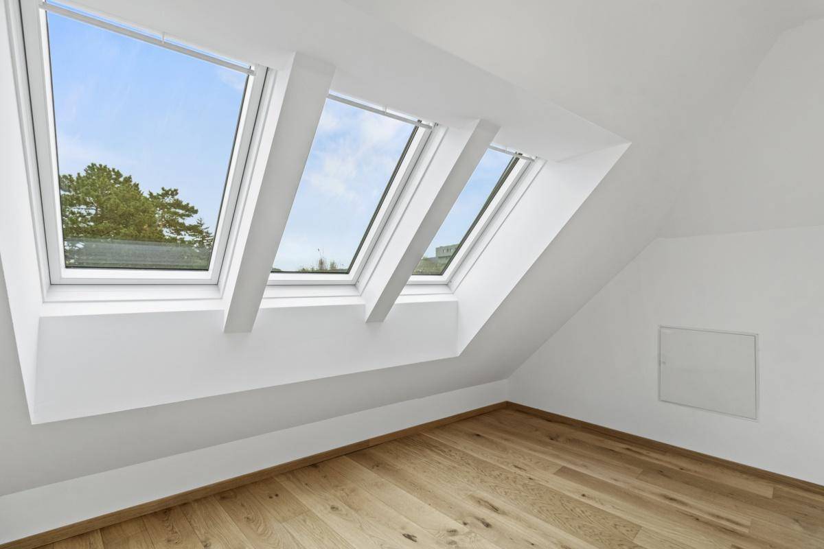 Helles Dachzimmer mit Holzboden und großen Dachfenstern, die viel Licht spenden und einen grünen Ausblick bieten.