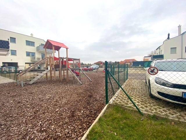 Kinderspielplatz mit Rutsche und Schaukeln, umgeben von einem Zaun und Parkplätzen.