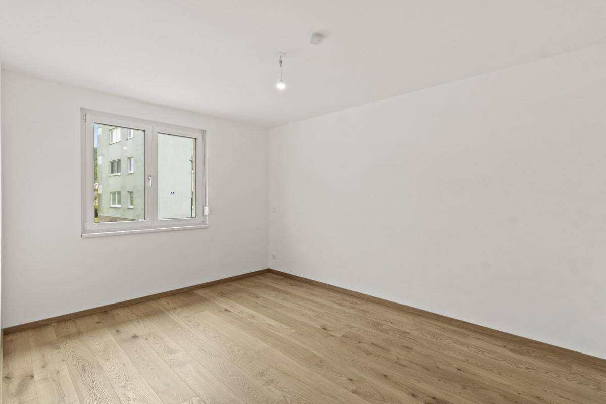 Helles Schlafzimmer mit Parkettboden und Blick auf ein gegenüberliegendes Gebäude durch das Fenster.