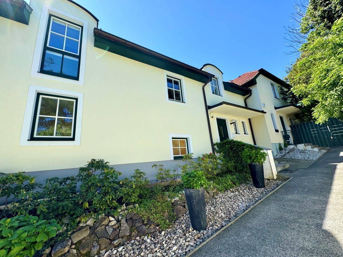 Seitenansicht des Hauses mit heller Fassade, grünen Fensterrahmen und gepflegter Bepflanzung im Vorgarten.
