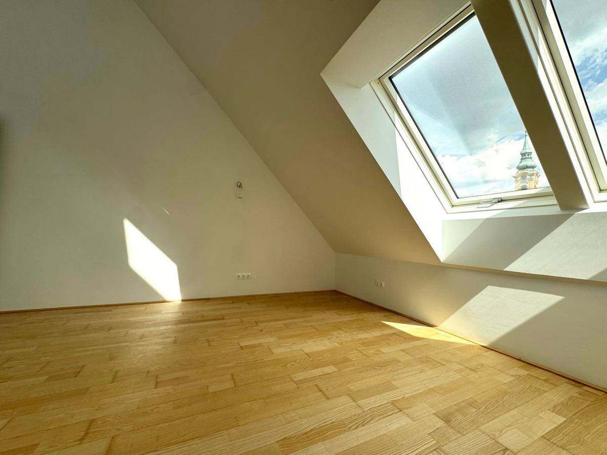 Heller Wohnbereich mit Holzparkettboden, schrägen Wänden und einem Dachfenster mit Blick auf die Stadt.