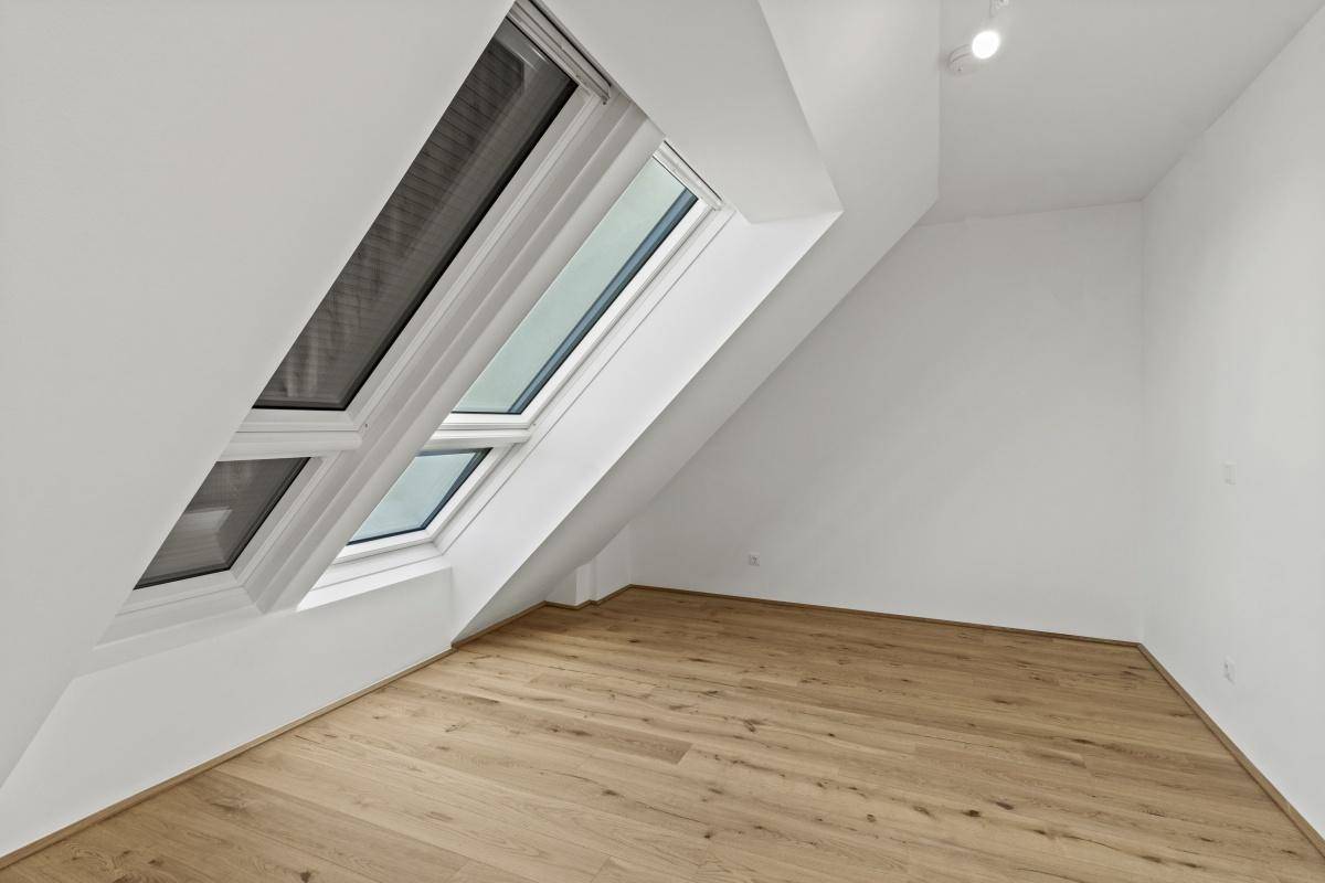 Heller Dachgeschossraum mit Holzboden und Dachfenstern, ausgestattet mit Jalousien für Sonnenschutz.