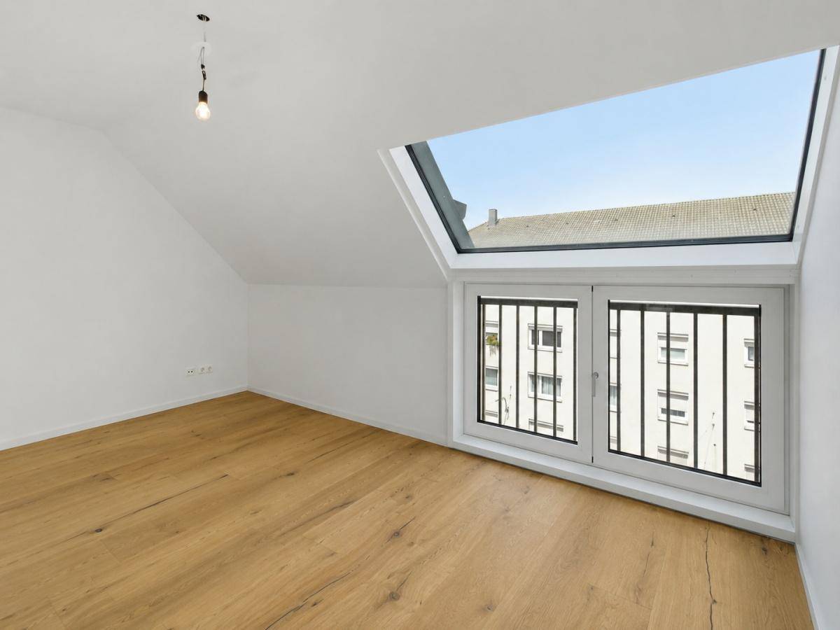 Geräumiger Dachgeschossraum mit Holzboden, weißen Wänden und einem großen Fenster mit Gitter.