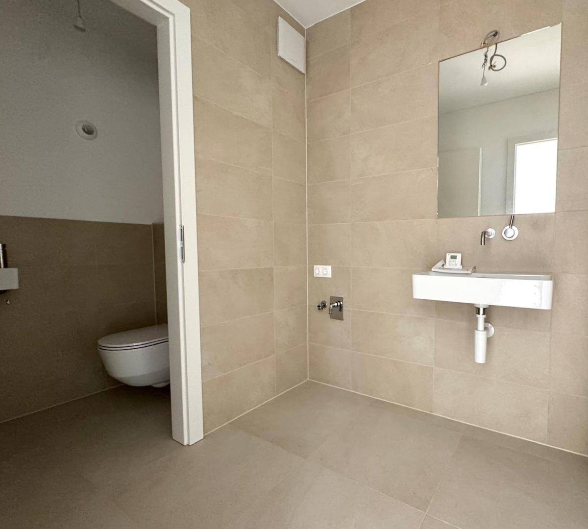 Modernes Badezimmer mit beigefarbenen Fliesen, wandhängender Toilette und einem eleganten Waschbecken.