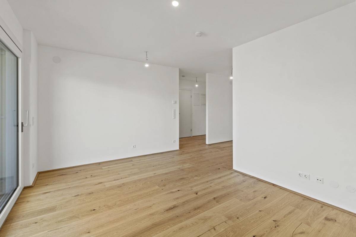Leeres, helles Wohnzimmer mit Holzboden und Zugang zu einem Balkon.