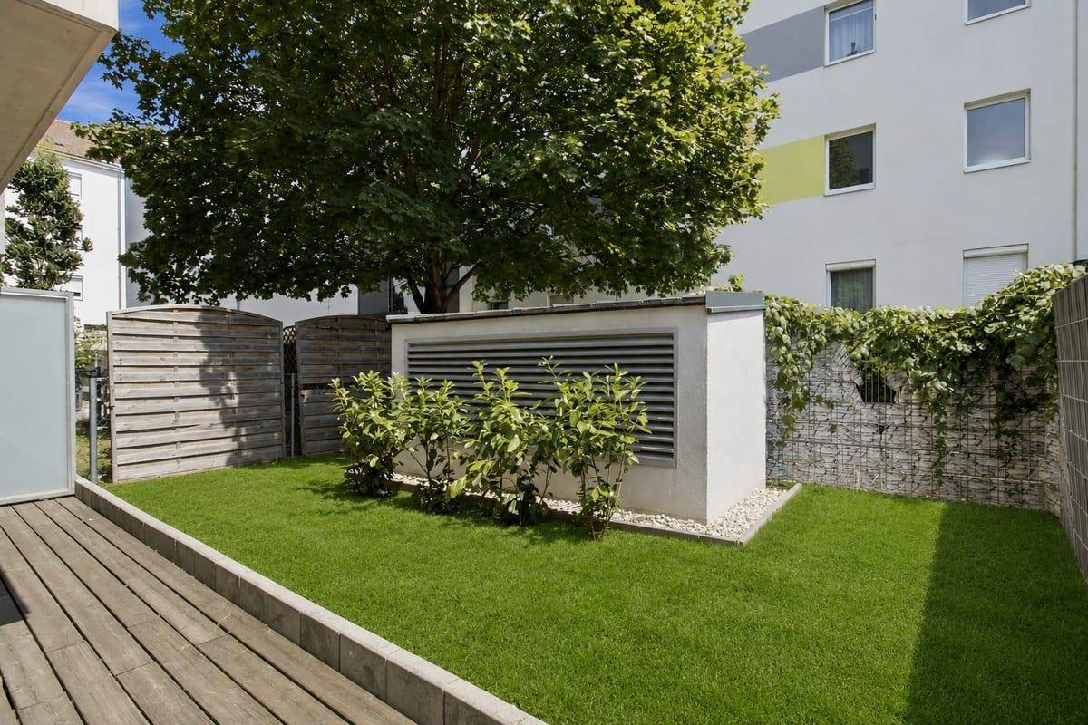 Ruhiger Gartenbereich mit Holzterrasse und Rasen, umgeben von einer Gabionenwand und Bepflanzung.